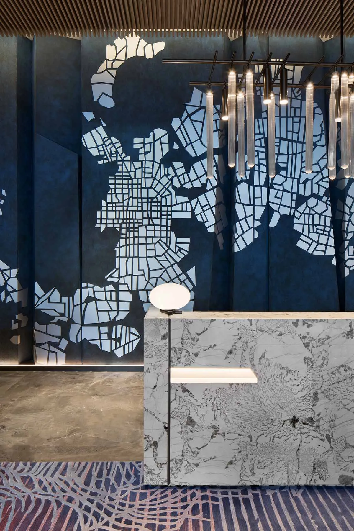 A modern lobby featuring a marble wall with a map pattern, ambient lighting, and geometric hanging lights.