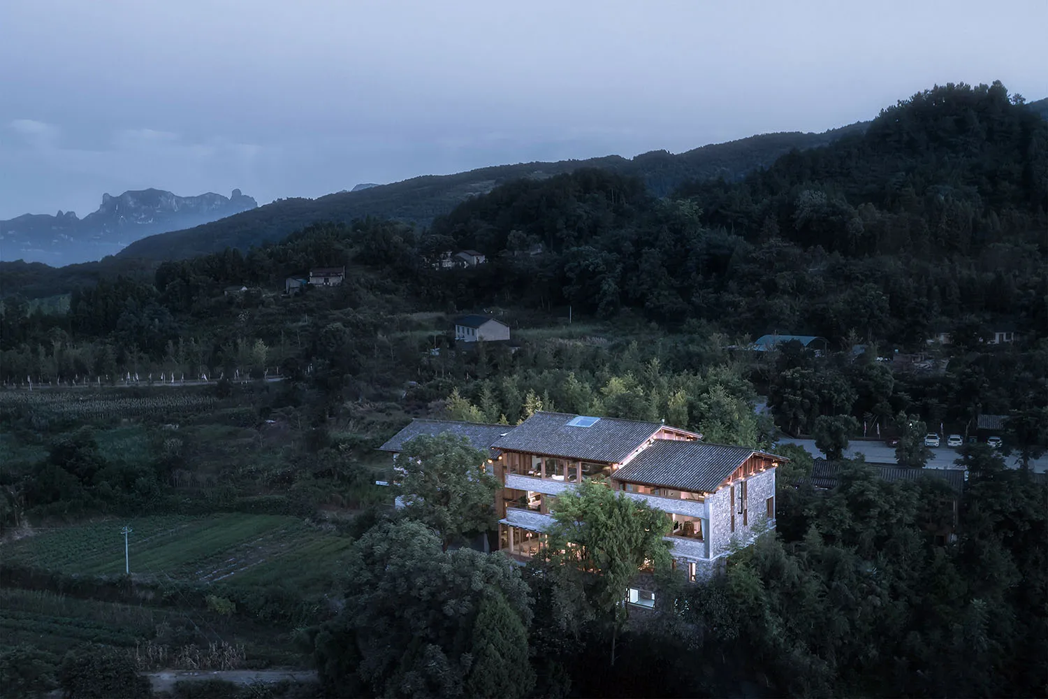 A modern residential building with a wooden roof and windows, surrounded by greenery and mountains.