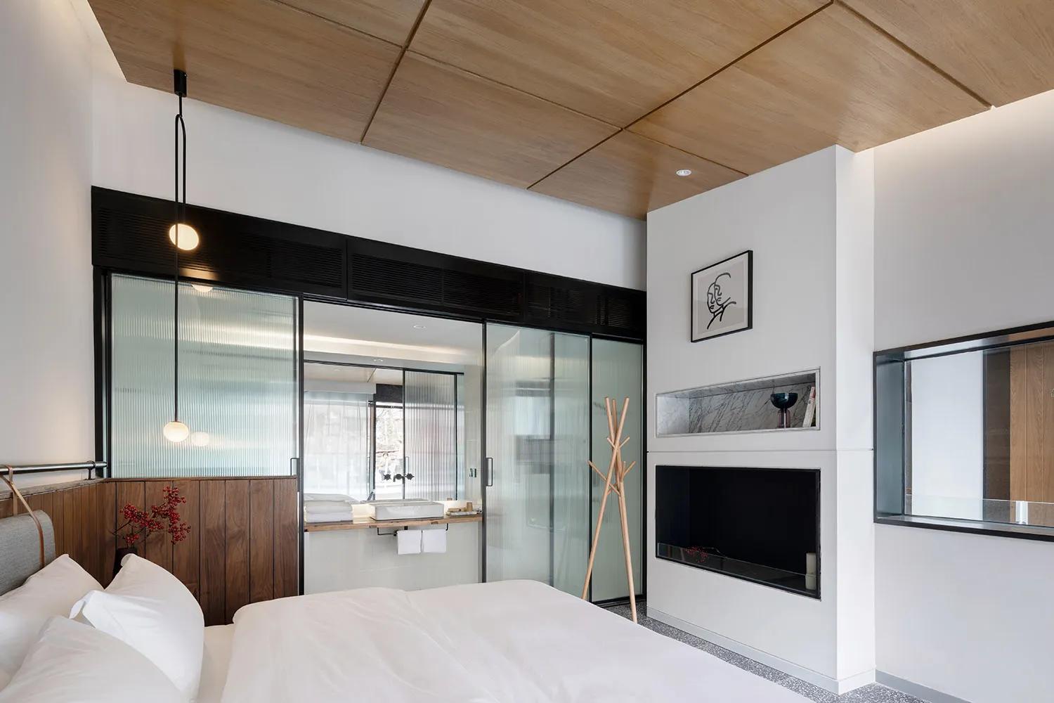 A minimalist bedroom featuring white bedding and lighting, with a glass-enclosed bathroom and wooden ceiling panels.