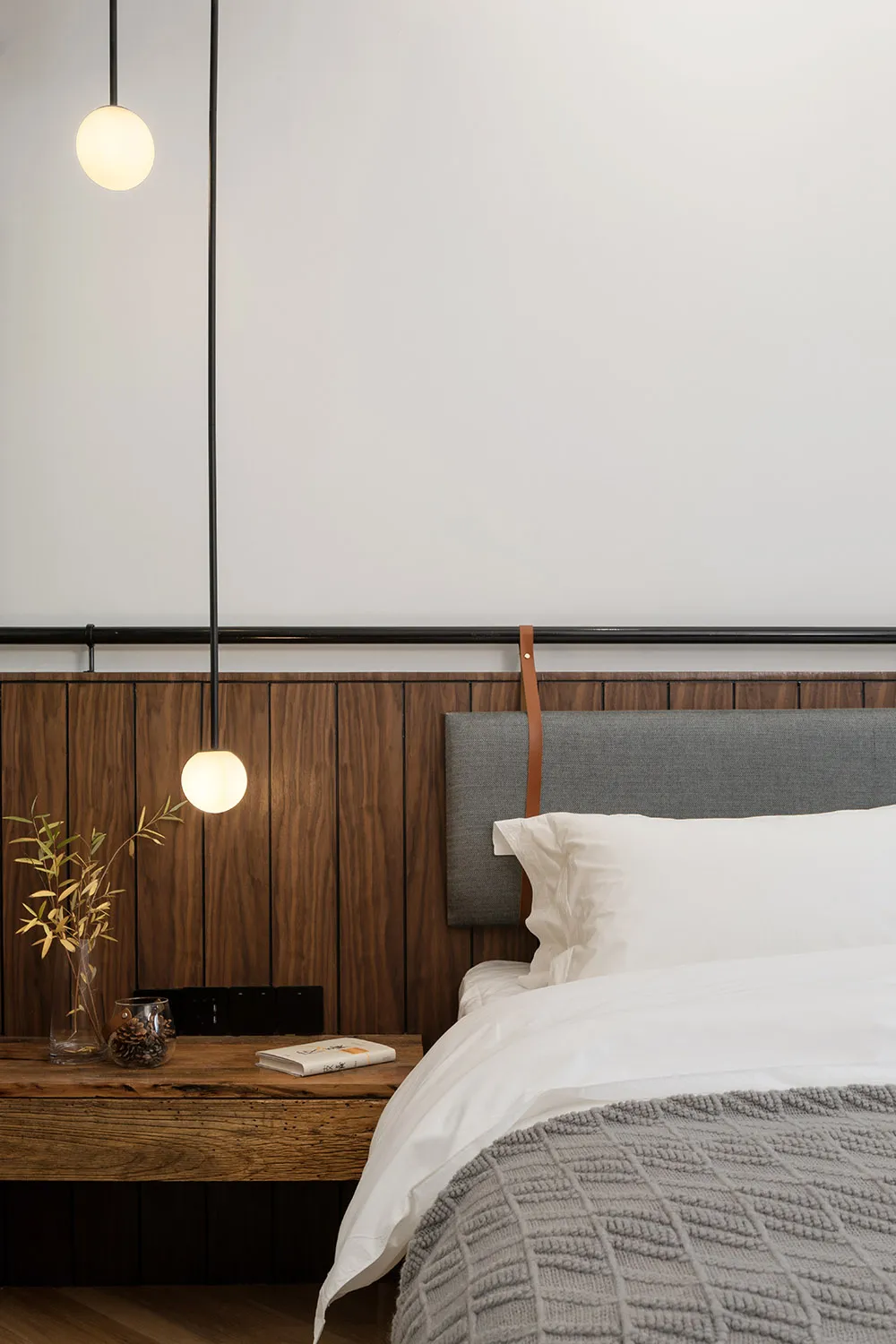 A minimalist bedroom featuring a wooden wall, a bed with a grey headboard, and a hanging light fixture.