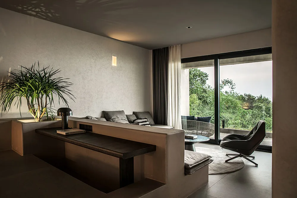 A minimalist living room with concrete walls, a gray sofa, and a black armchair, featuring a large window with a view of gree