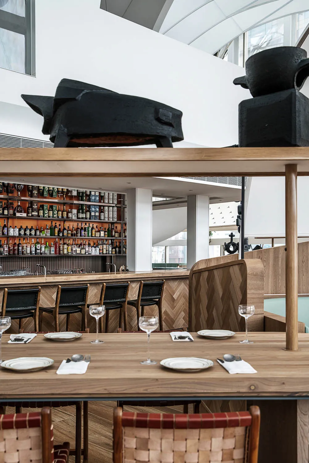 Modern restaurant interior with wooden tables, metal stools, and glass shelves displaying liquor.