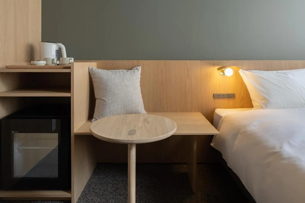 A minimalist bedroom featuring a bed with wooden headboard and nightstand, a small wooden side table, and a wall-mounted lamp