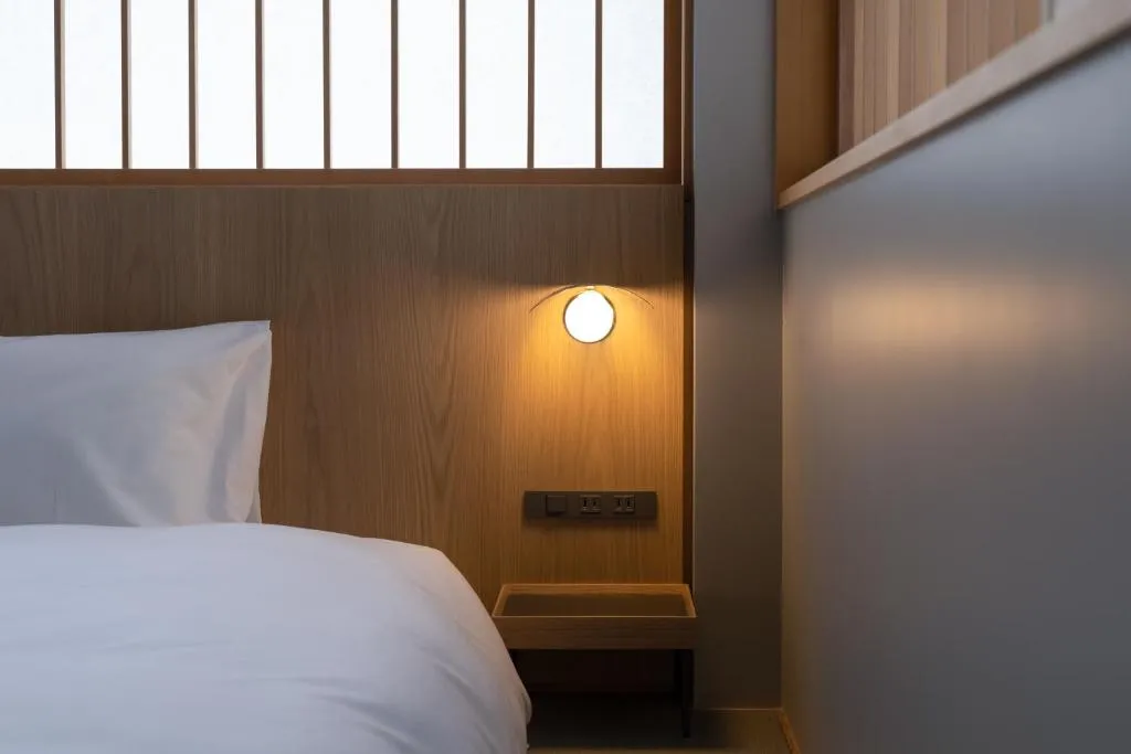 A minimalist bedroom featuring a white bed with a wooden headboard and light fixture against a wall of light grey flat panels