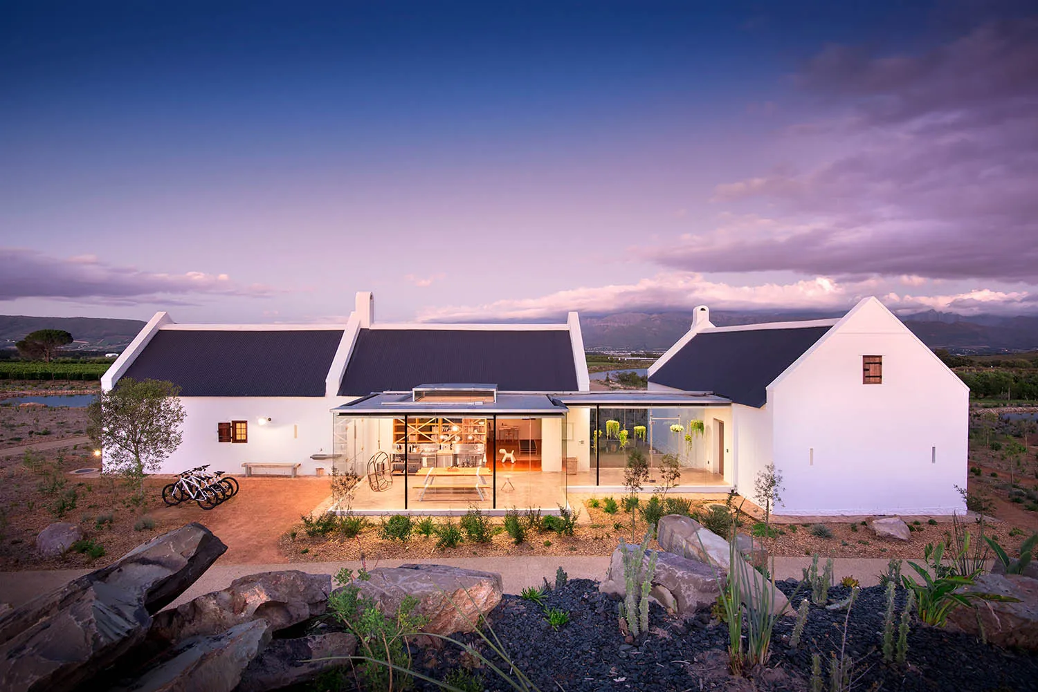 White minimalist house with concrete walls and large glass windows, set against a backdrop of mountains and clouds.