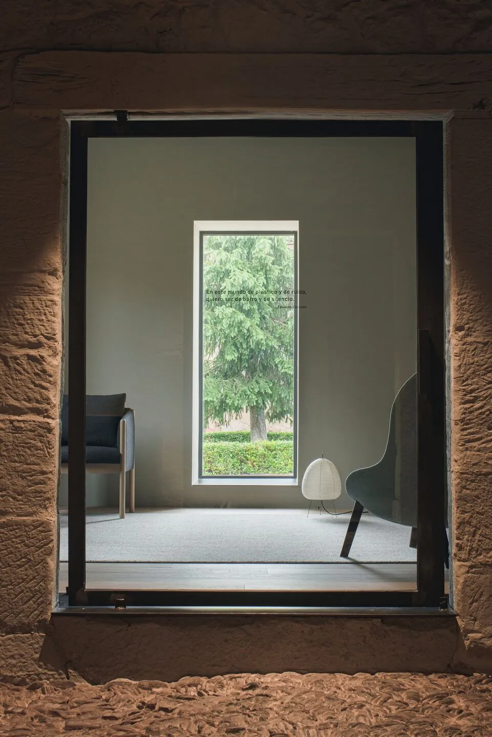 Modern living room with concrete walls and floor, large window displaying greenery, minimalist furniture, and a floor lamp.