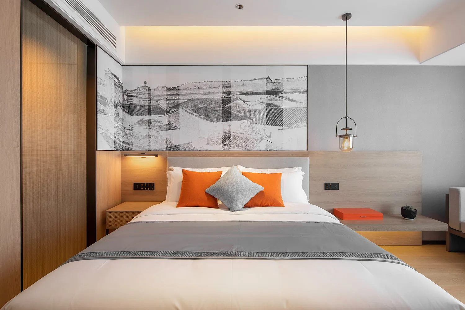 A stylish, minimalist bedroom featuring a bed with orange cushions and a gray blanket, complemented by a framed black and whi