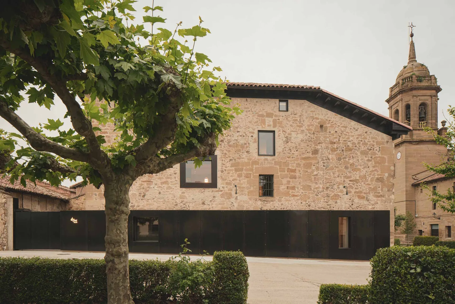 The exterior of a modern building made of stone with large windows and a tree in the foreground.