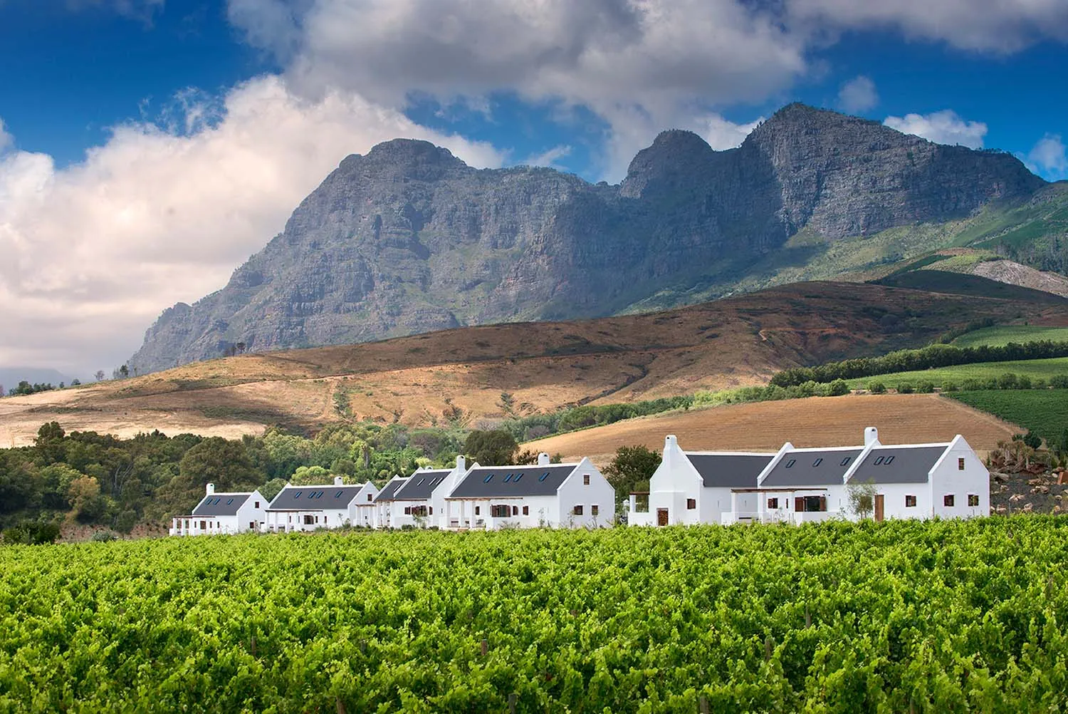 A picturesque village with white tiled rooftops surrounded by lush green fields and dramatic mountains in the background.