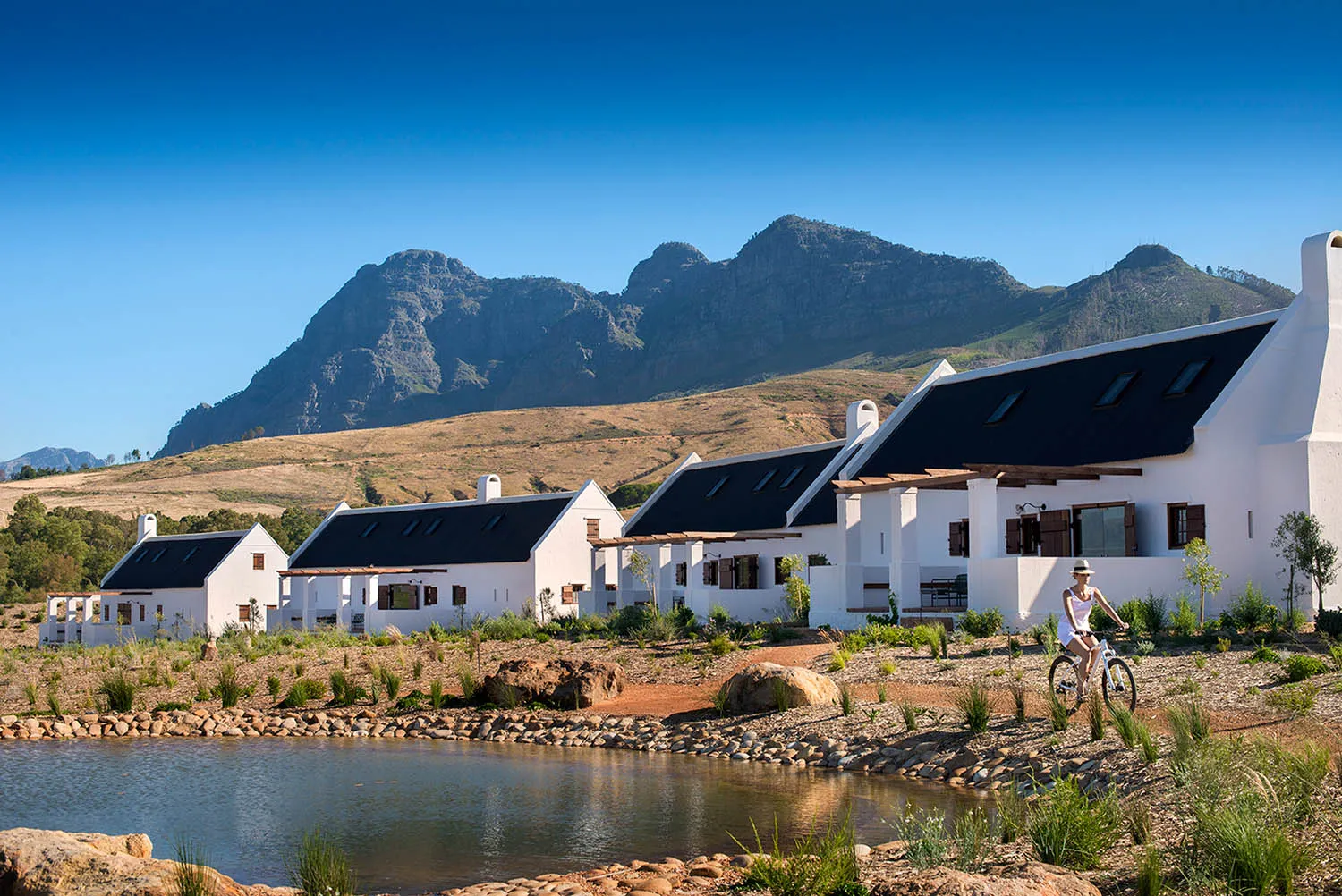 A row of white stucco residential villas surrounded by a small pond and a scenic mountain backdrop.