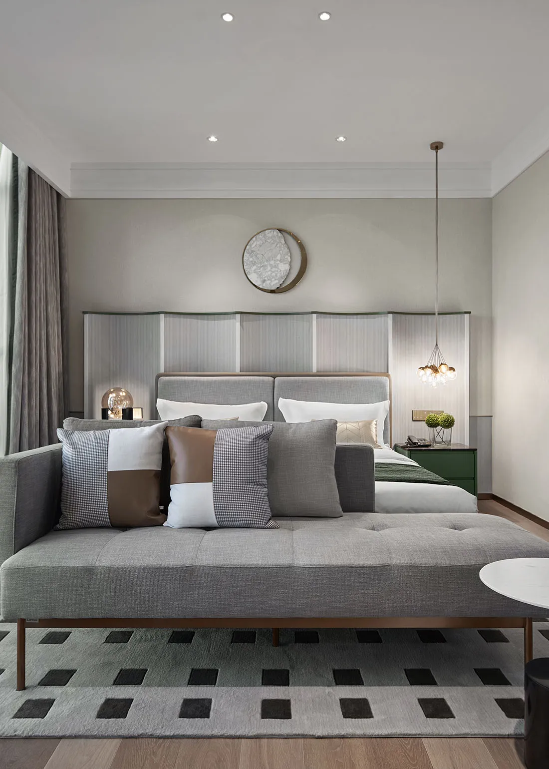 A minimalist bedroom featuring a grey fabric bed with multiple cushions, a green nightstand, and a round ceiling light.
