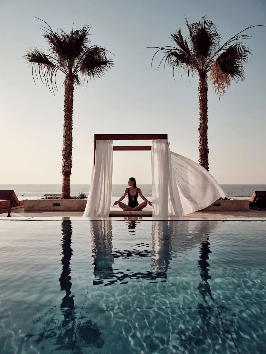 A woman in a minimalist setting sitting on a sun drape by an infinity pool with tropical surroundings