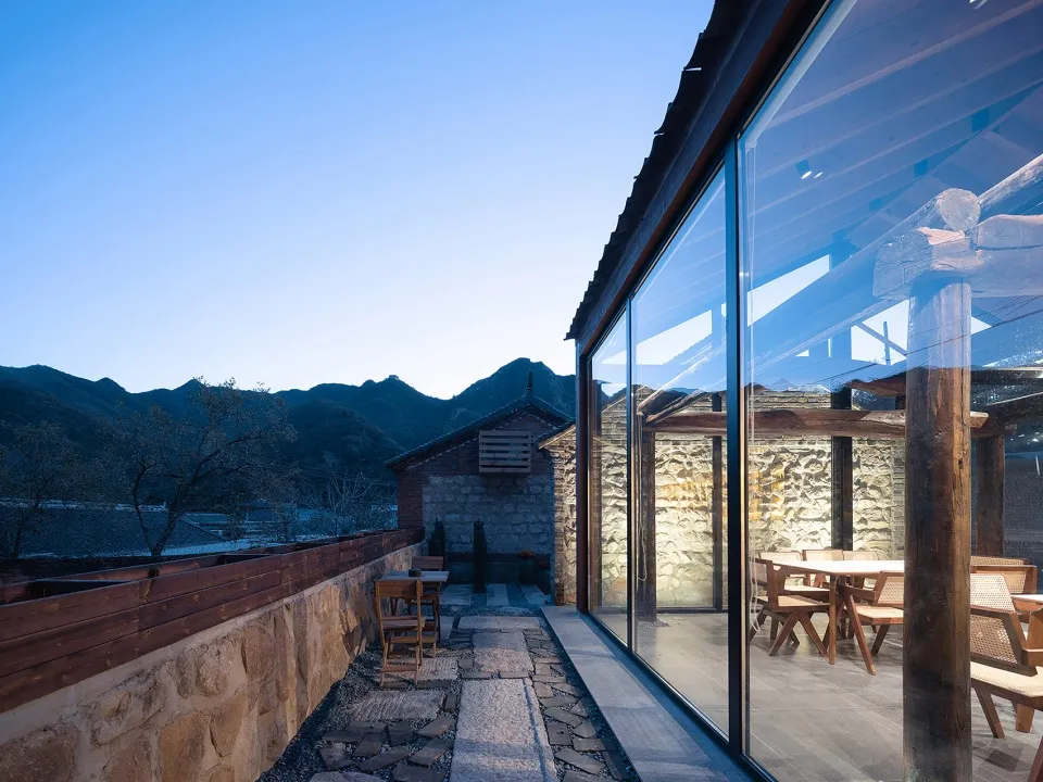 Outdoor seating area with stone walls, glass walls, and concrete patio at dusk, overlooking mountains.