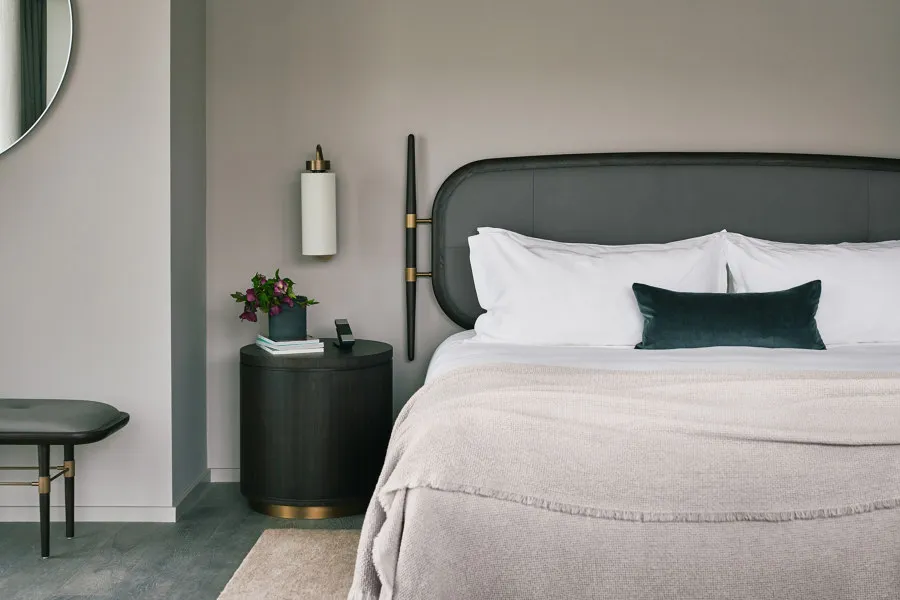 A modern bedroom featuring a fabric bed with a black headboard on a grey floor, accompanied by a side table with a lamp and a