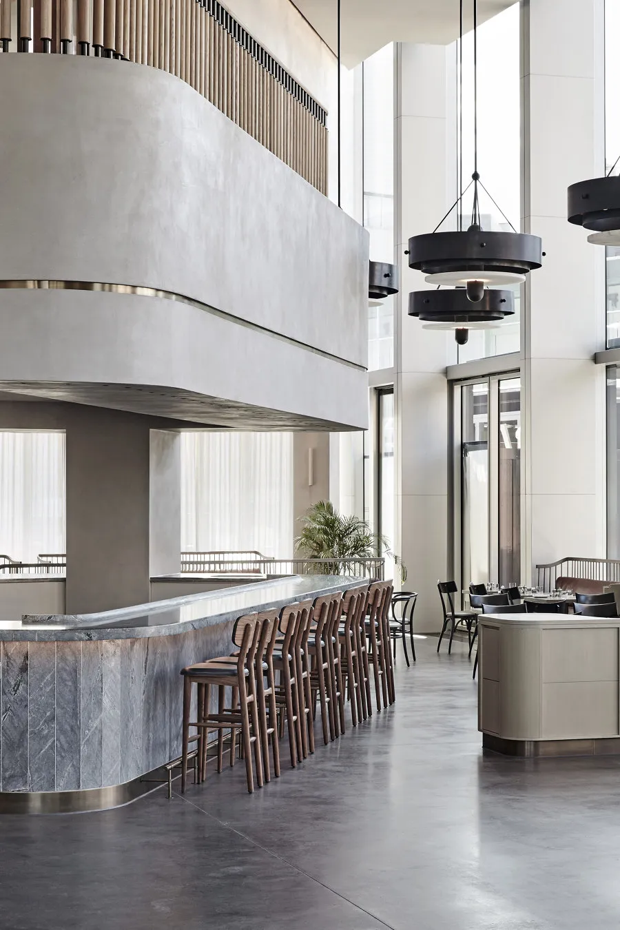 A modern industrial restaurant bar with concrete and marble surfaces, featuring wooden chairs and black pendant lights.