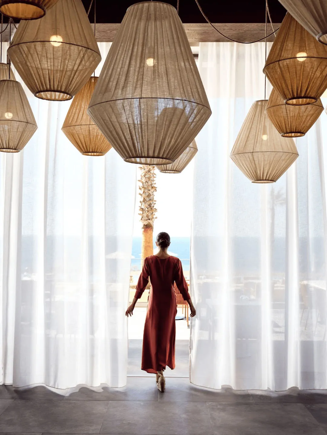 A woman in a red dress walks through white curtains towards the ocean, surrounded by hanging fabric lanterns.
