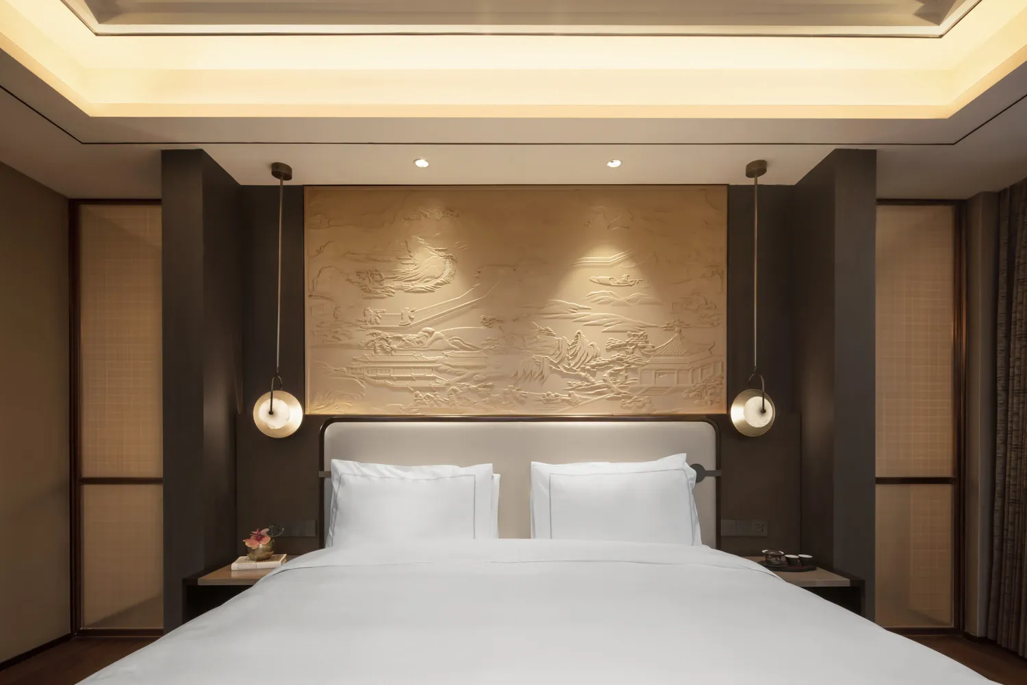 A modern minimalist bedroom interior with white bedding and patterns, featuring a decorative headboard and lighting fixtures.
