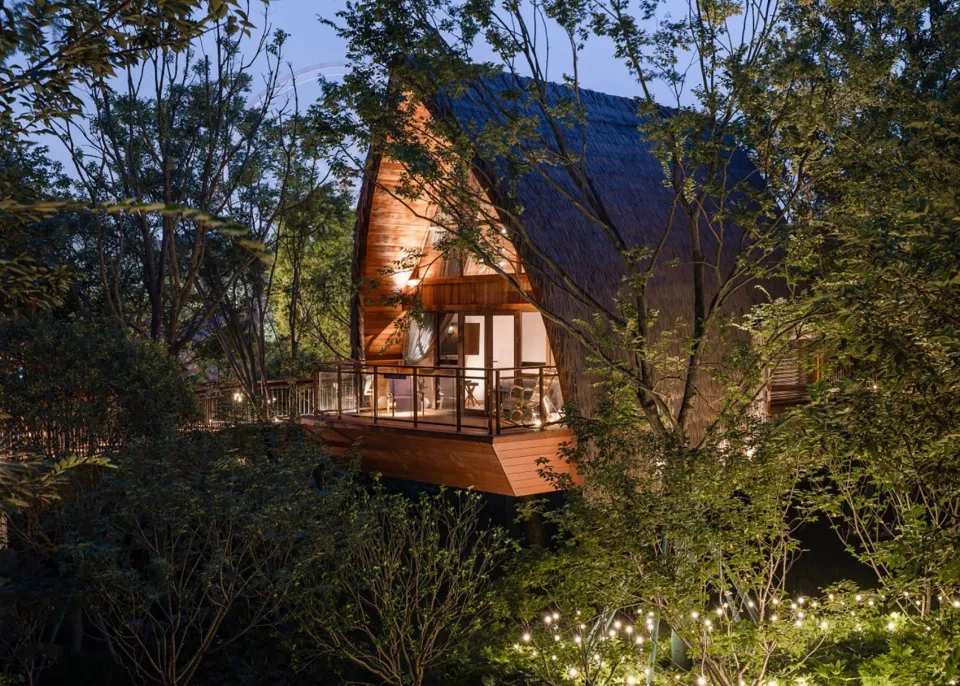 A modern wooden treehouse with lights illuminated at night, surrounded by trees.