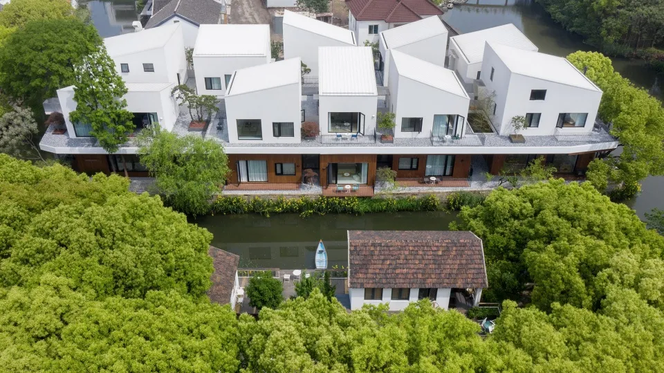 Aerial view of modern black-roof houses with white stucco exteriors and geometric designs, surrounded by greenery and water.