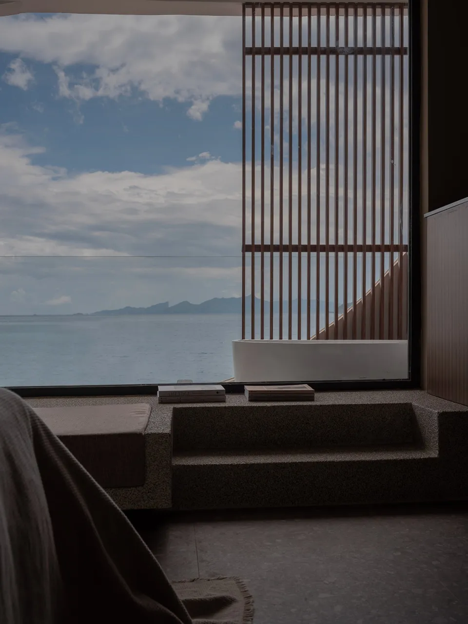 極簡度假房間,石材包覆與木質牆板,面海景,配有浴缸與書籍。