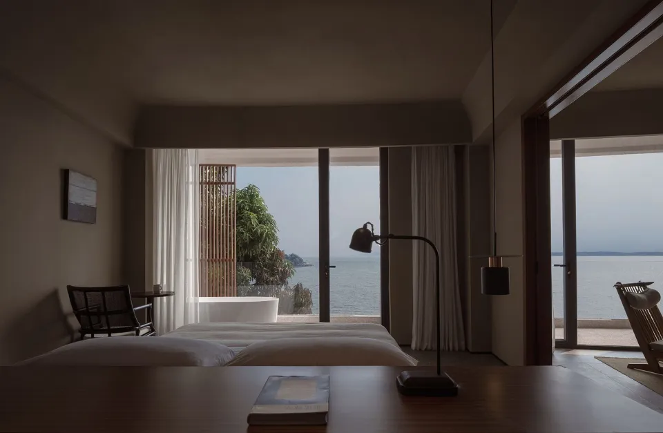 Modern minimalist bedroom with wooden furniture, fabric bedding, and metallic accents overlooking the ocean.