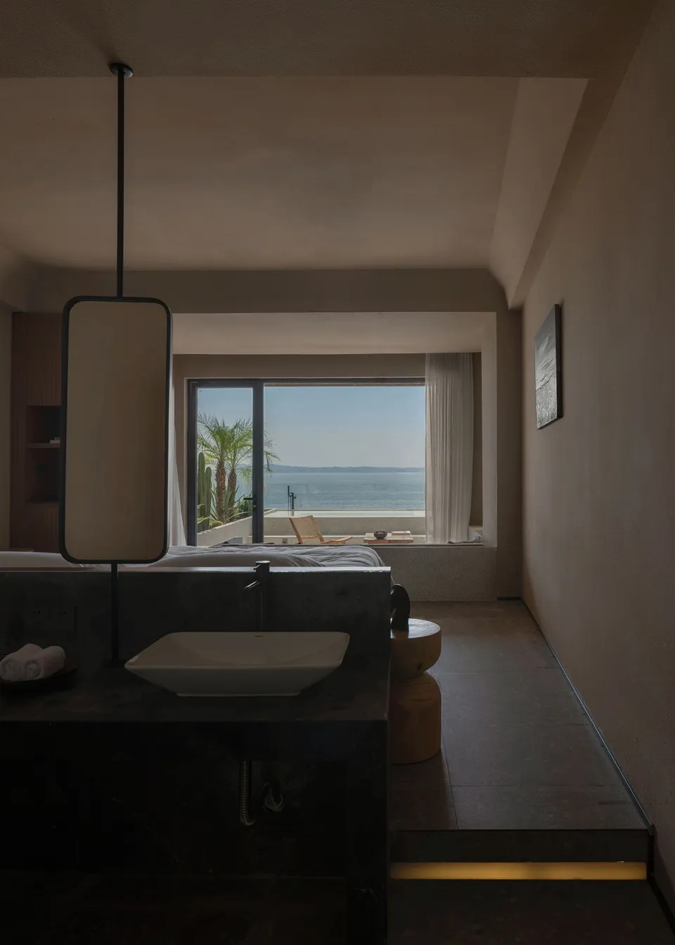 Modern bedroom with concrete walls and floor, large window overlooking the ocean, and minimalist furniture.