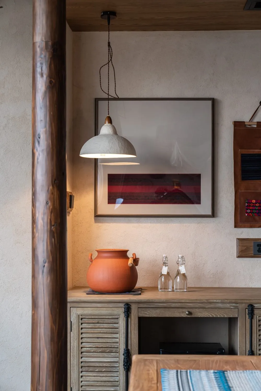 A modern living room with wooden furniture, featuring a pendant light, a red and white framed picture, and a terracotta vase.