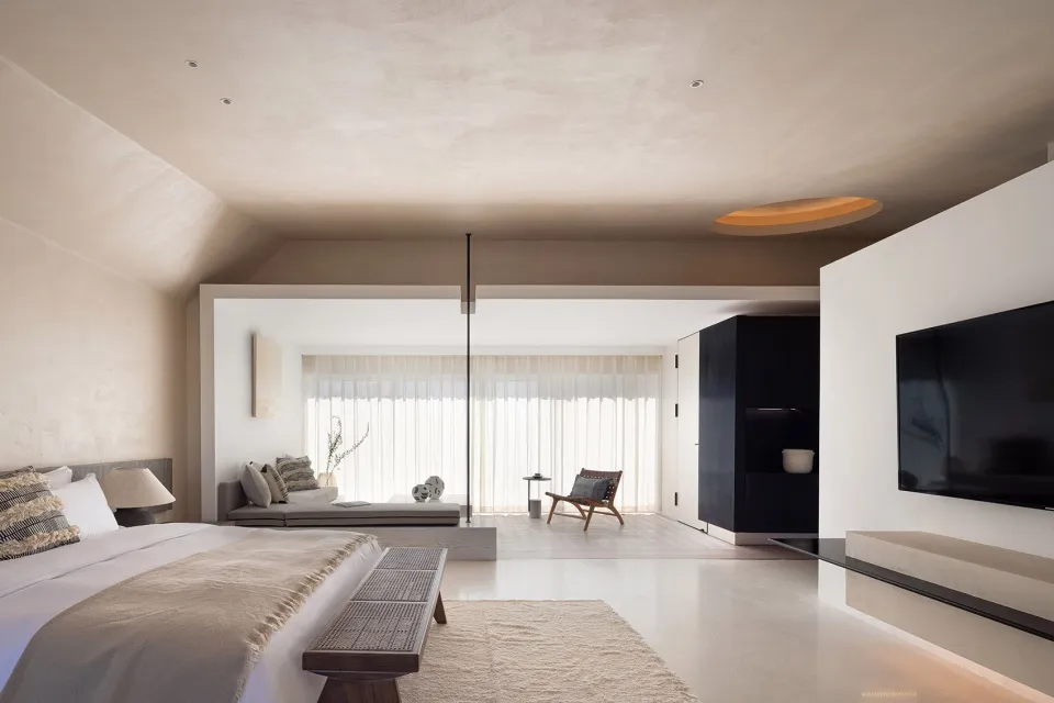 Modern minimalist bedroom with neutral materials, featuring a large bed, a sofa, and a TV.