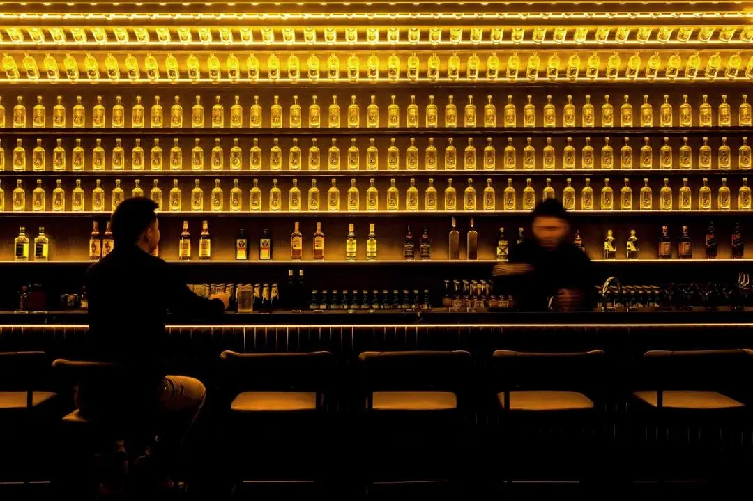 A modern bar interior with a wall of illuminated liquor bottles.
