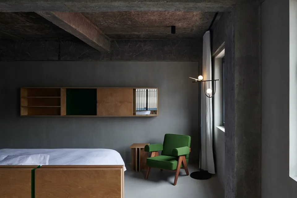 Minimalist bedroom with concrete walls, wooden furniture, and a green upholstered chair.