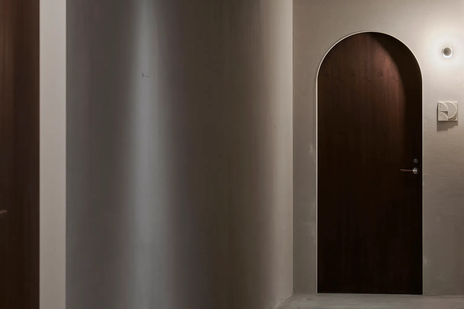 A modern, arched wooden doorway against a white wall with a light fixture above it.