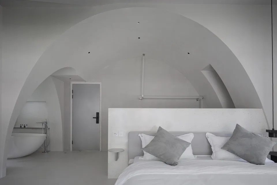 A minimalist bedroom with concrete walls, a white bed and pillows, and a sleek white bathtub in the corner.