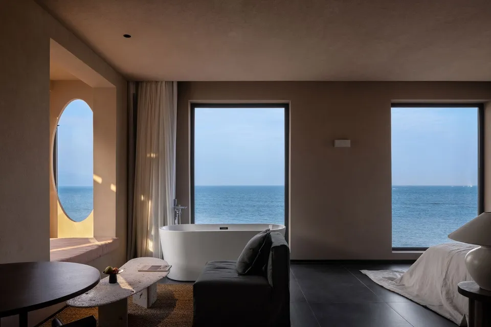 Modern hotel suite featuring a concrete bathtub, glass windows overlooking the ocean, and a cozy seating area with a sofa and