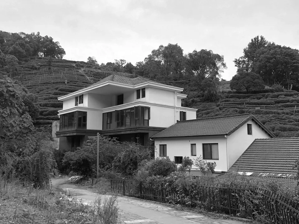 A modern residential house with a concrete exterior, surrounded by greenery and a hillside covered in terraced fields.