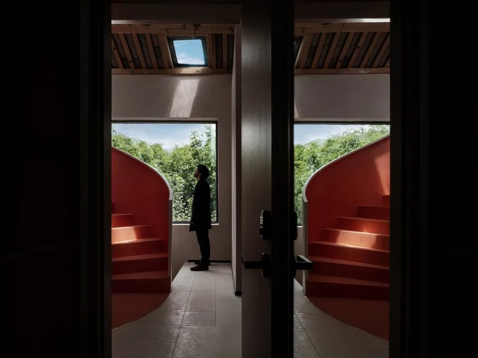 A person standing in a modern residential space with red accents and windows overlooking greenery.