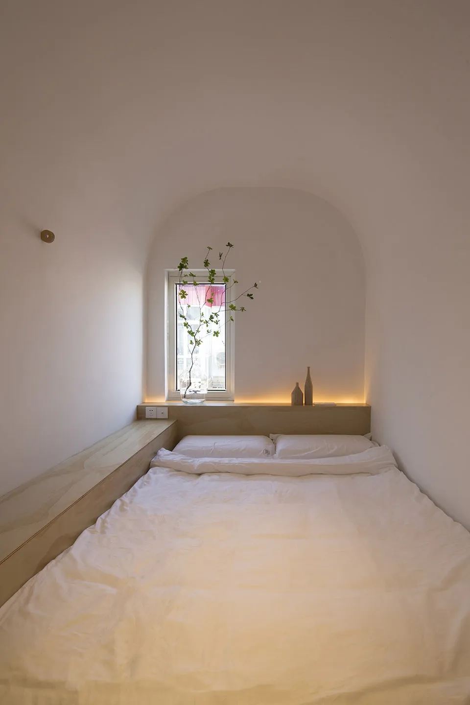 A minimalist bedroom with a white bed and wooden frame, featuring a small window with a plant and two decorative items on the