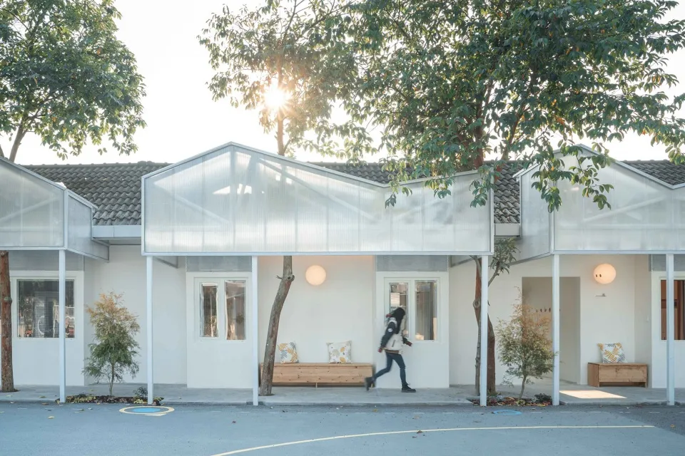 A modern house with glass walls and wooden elements sits beneath tall trees.
