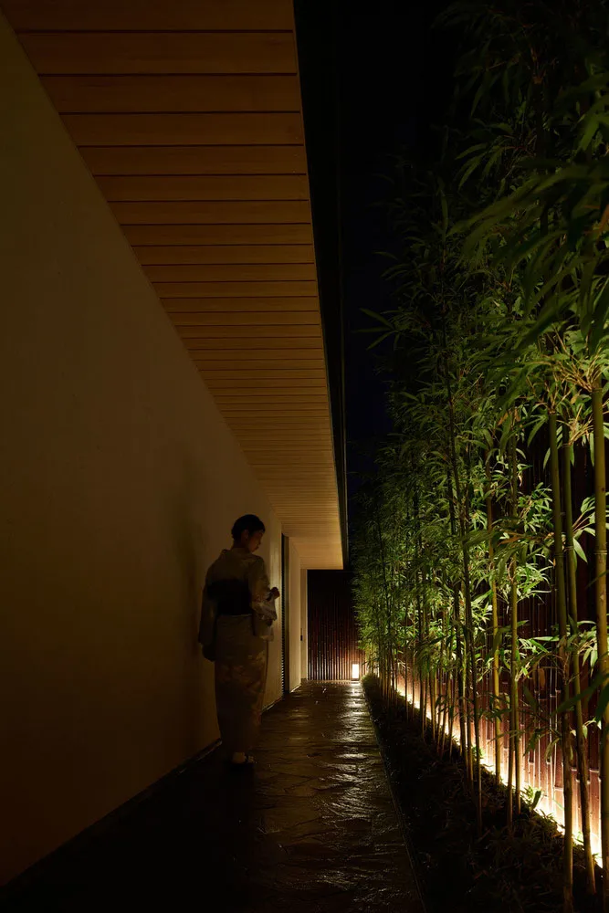 A woman in a kimono standing on a stone path bordered by tall bamboo plants illuminated by lights.