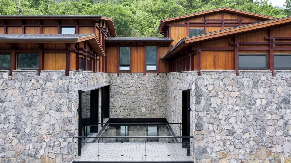 Modern residential building with wood and stone exterior, featuring large windows and a stone wall with a glass railing.
