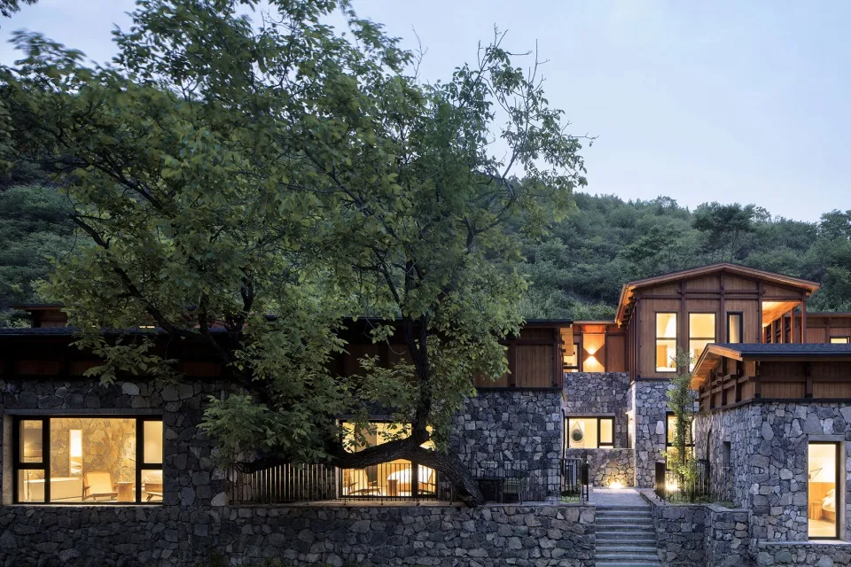 A modern residential house with stone walls, wooden structures, and large windows, surrounded by greenery.