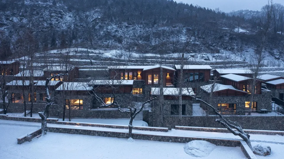 A modern residential architecture nestled in a snowy valley with stone and glass materials.