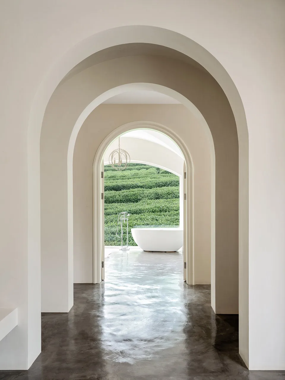Modern minimalist bathroom featuring a white bathtub on a polished concrete floor, framed by arches of plaster and viewed thr