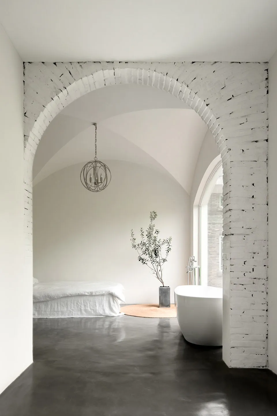 Modern minimalist bedroom with a white bed and a round tree in a vase next to a freestanding bathtub, all under a white brick