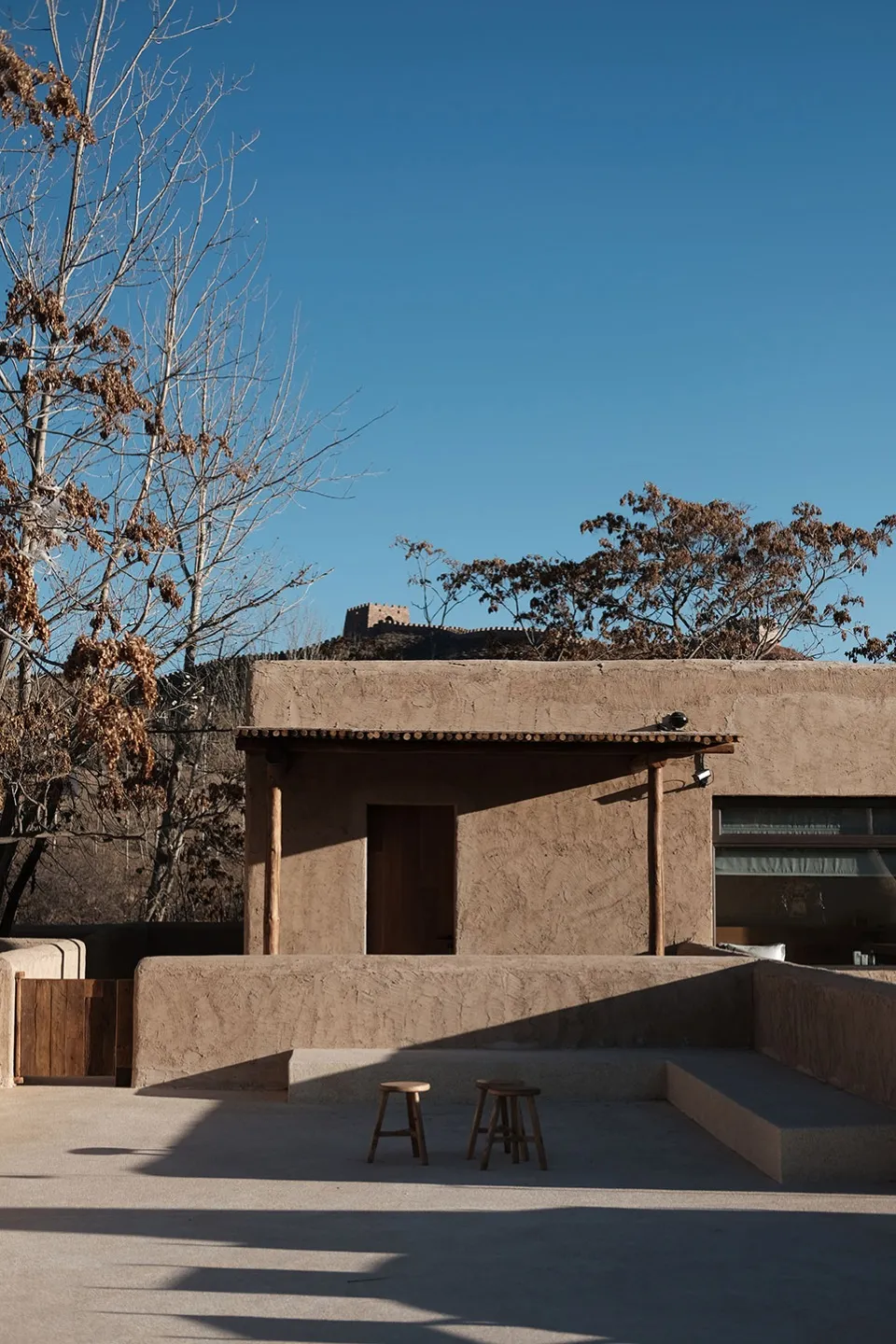 A minimalist clay exterior building with a wooden door and two stools in front of it.