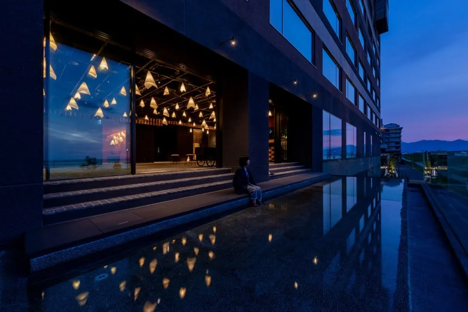A modern hotel exterior with glass walls and roof lights reflecting in the water below.