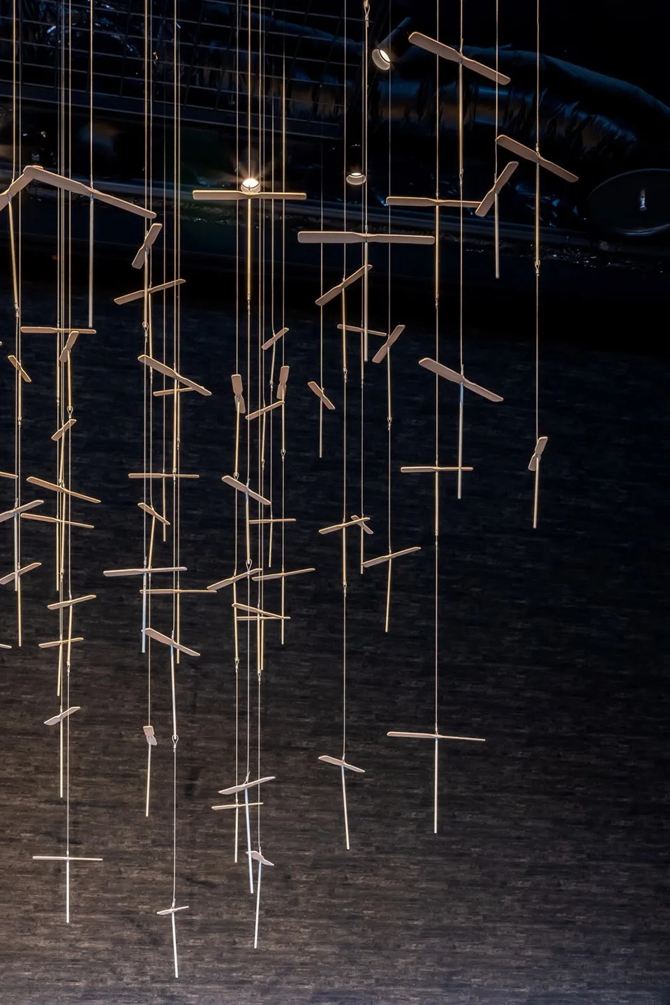 The image depicts a modern installation featuring geometric wood sculptures with attached metal rods illuminated by hanging l