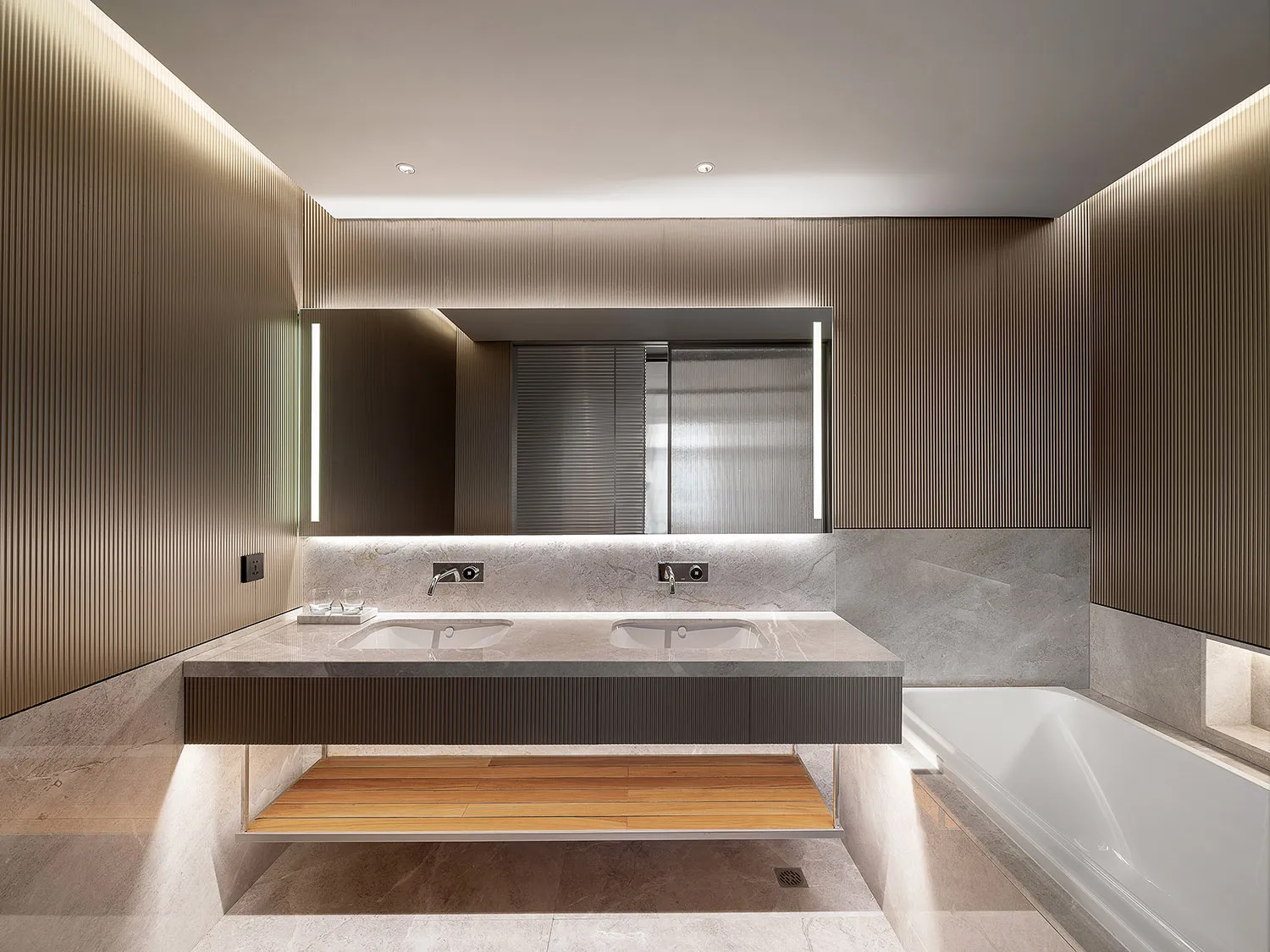 A modern bathroom features a marble vanity with concrete accents and a wooden shelf beneath it, illuminated by soft lighting.