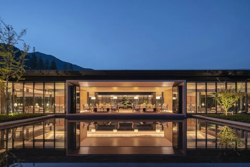 Modern residential house exterior with glass windows, concrete elements, and warm lighting at dusk.