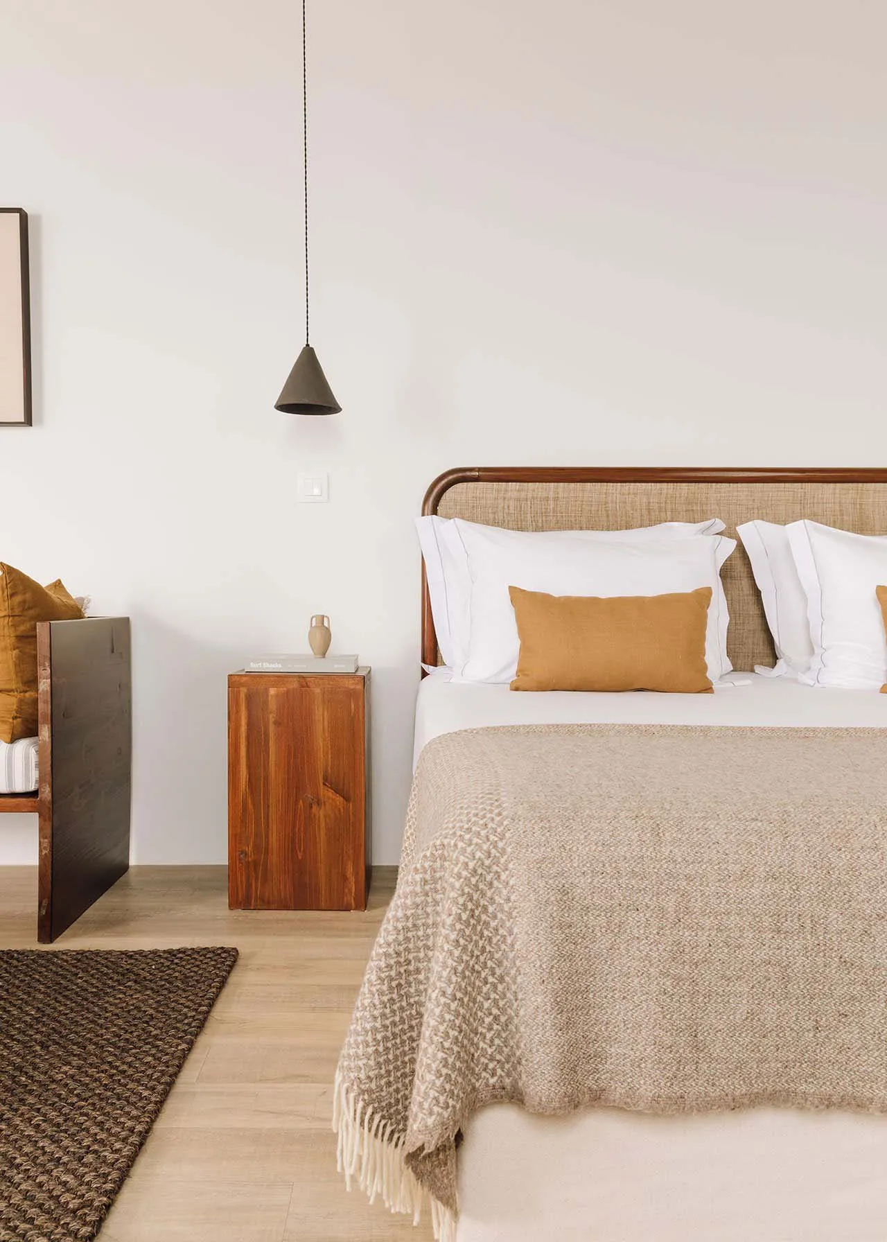 A minimalist bedroom featuring a white bed with a beige textured blanket, white pillows, and a wooden nightstand with a brown