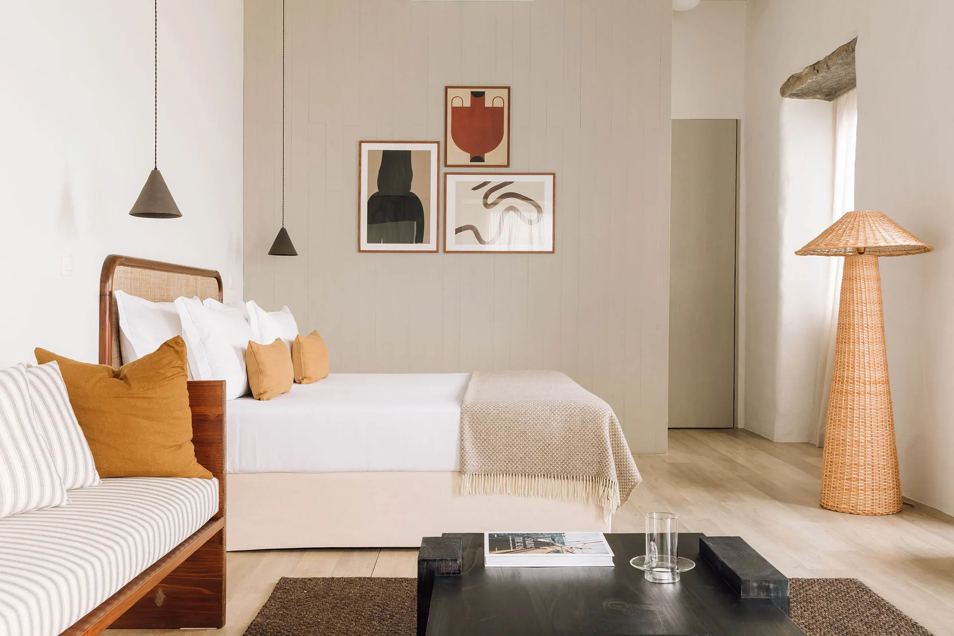 A minimalist bedroom with neutral tones, featuring a bed, modern pendant lights, abstract art, and a rattan floor lamp.
